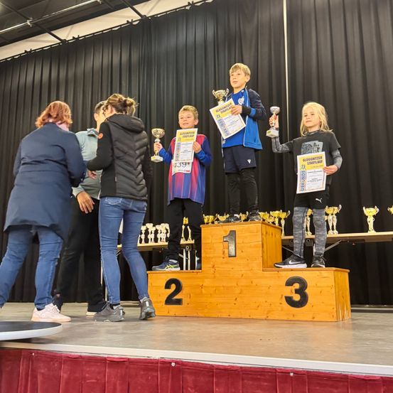 Eine Gruppe junger Athleten steht mit Trophäen auf dem Podium, einer hält ein Schild, während eine Frau weitere Trophäen verteilt.