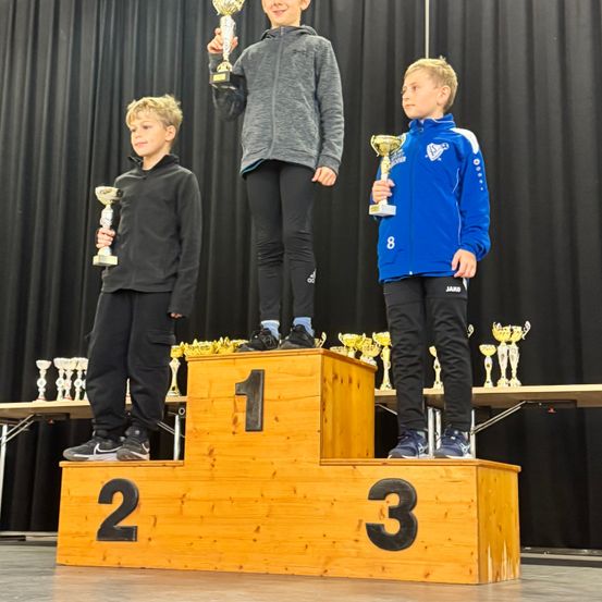 Drei Jungen stehen auf einem Podest mit den Nummern 1, 2 und 3. Der Junge in der Mitte hält einen Pokal. Hinter ihnen steht ein Tisch mit Pokalen. Der Junge auf der linken Seite trägt eine schwarze Jacke und Hose. Der Junge auf der rechten Seite trägt eine blaue Jacke und Hose.