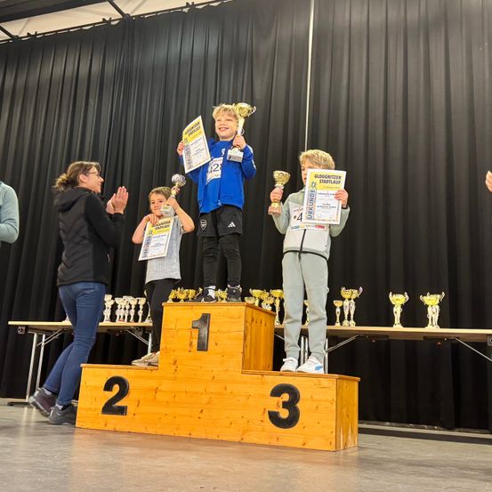 Drei Kinder stehen auf einem Podest und halten Trophäen. Das Kind links hält ein Zertifikat mit der Nummer 22. Das mittlere Kind hält eine Trophäe mit der Nummer 42. Das Kind rechts hält ein Zertifikat mit der Nummer 4.