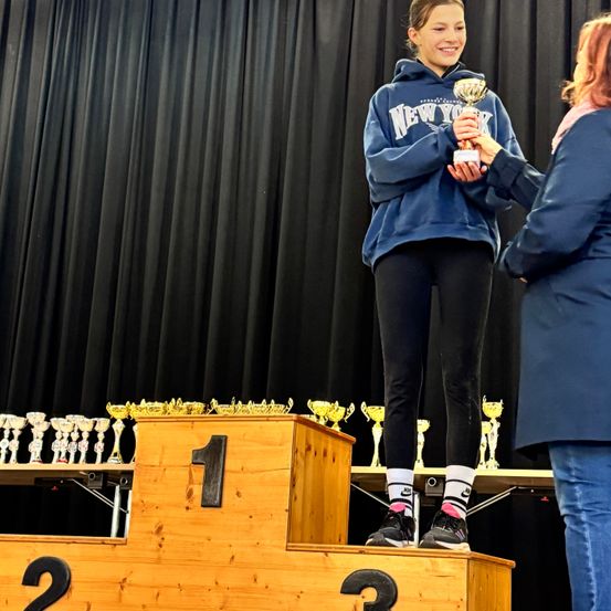 Ein Mädchen in einer blauen Kapuzenjacke steht auf dem Podest des ersten Platzes und hält eine Trophäe. Eine Frau in einem blauen Mantel steht neben ihr. Dahinter ist ein schwarzer Vorhang und mehrere Trophäen zu sehen.
