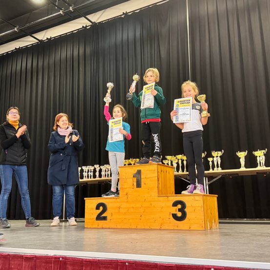 Drei Kinder stehen mit Trophäen auf einem Podest, während zwei Erwachsene hinter ihnen stehen. Sie sind auf einer Bühne, und es gibt mehr Trophäen und Pokale auf einem Tisch im Hintergrund.
