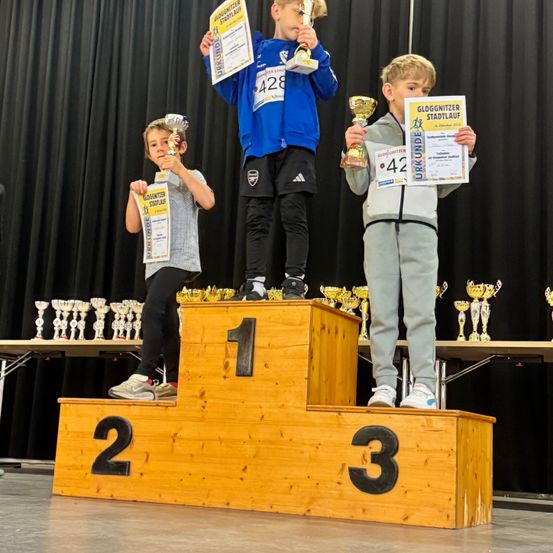 Drei Kinder stehen auf einem Podest mit Trophäen. Ein Kind hält eine Trophäe, ein anderes hält zwei und das dritte Kind hält drei Trophäen. Sie stehen auf einem hölzernen Podest mit den Nummern 1, 2 und 3.