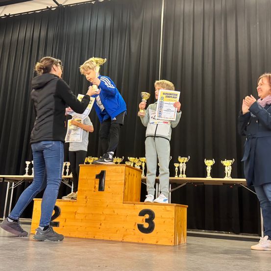 Zwei junge Jungen stehen auf einem Podium mit Trophäen. Eine Frau übergibt einem Jungen eine Trophäe. Ein anderer Junge hält ein Zertifikat. Ein Tisch mit Trophäen steht hinter ihnen.