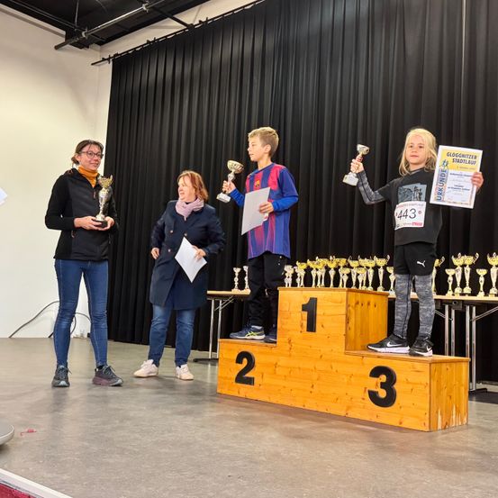 Vier Personen stehen auf einer Bühne, zwei halten Trophäen, während zwei präsentieren. Dahinter sind Trophäen auf einem Tisch ausgestellt. Die Bühne hat ein Podest mit den Zahlen 1, 2 und 3.