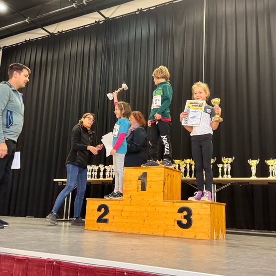 Drei junge Athleten stehen auf einer Bühne mit Podesten hinter ihnen. Sie halten Pokale und Urkunden. Die Bühne ist mit schwarzen Vorhängen und einem roten Teppich geschmückt.