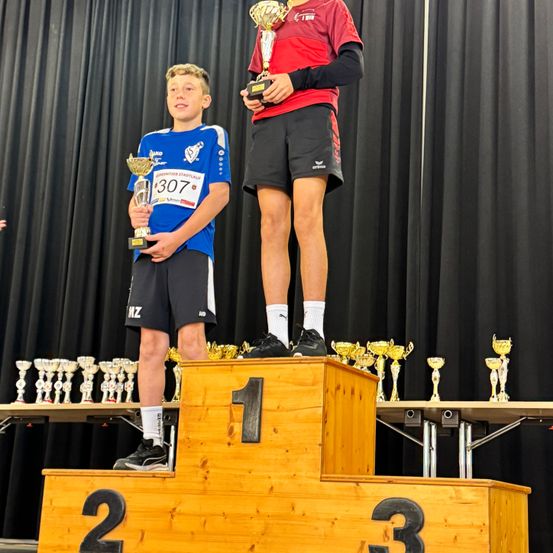 Zwei junge Athleten stehen auf einem Podest, wobei der in Rot eine Medaille um den Hals trägt. Der andere hält eine Trophäe und trägt ein blaues Trikot. Trophäen stehen hinter ihnen auf einem Tisch.