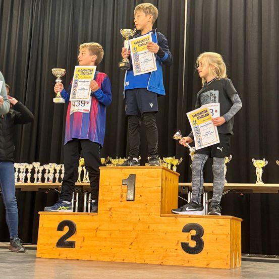 Drei Kinder stehen auf einem Podest mit Trophäen, tragen Turnschuhe und halten Urkunden. Sie haben die Nummern 1, 2 und 3 auf dem Podest. Dahinter stehen Trophäen auf einem Tisch.