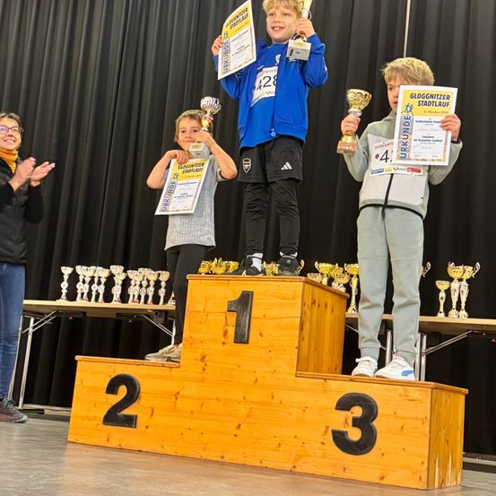 Drei Kinder stehen auf einem Podium mit einer Trophäe. Sie tragen Medaillen und halten Urkunden. Das Podium hat die Zahlen eins, zwei und drei. Dahinter steht ein schwarzer Vorhang.