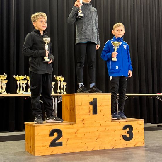 Drei junge Jungen stehen mit Trophäen auf einem Podium, jeder eine in jeder Hand. Der Junge in der Mitte steht auf der obersten Stufe, während die anderen beiden auf den unteren Stufen stehen. Hinter ihnen befindet sich ein schwarzer Vorhang und ein Tisch mit Trophäen.