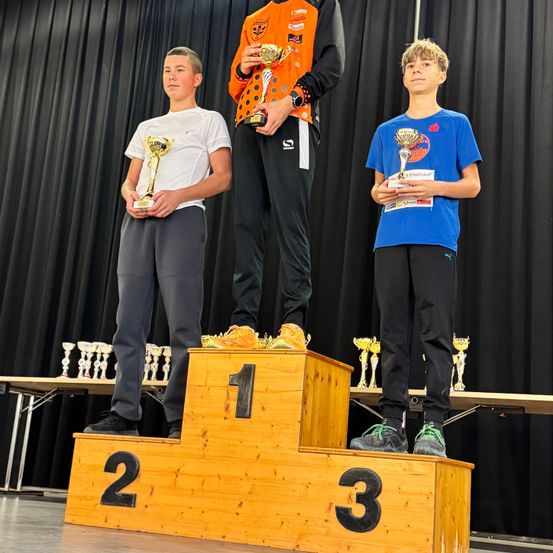 Drei Jungen stehen auf einem Podium, einer in der Mitte trägt eine orange-schwarze Jacke und hält zwei Trophäen. Der Junge links trägt ein weißes T-Shirt und hält eine Trophäe. Der Junge rechts trägt ein blaues T-Shirt und hält eine Trophäe. Hinter ihnen stehen Trophäen auf einem Tisch.