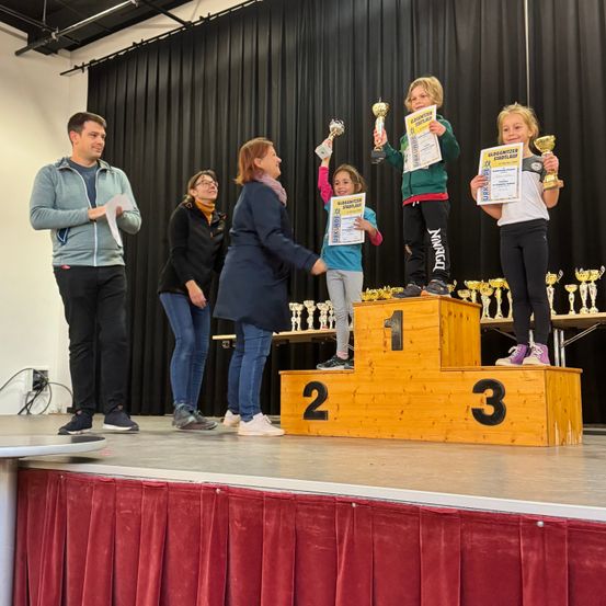 Eine Gruppe von Kindern steht auf einem Podium mit Trophäen. Sie werden von Erwachsenen begleitet, von denen einer ein Papier hält. Dahinter sind Trophäen auf einem Tisch ausgestellt. Die Umgebung ist auf einer Bühne mit Vorhanghintergrund.