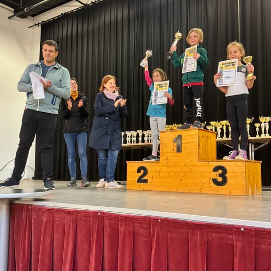 Ein Podium mit drei Kindern, die darauf stehen und Trophäen halten. Ein Mann steht links und hält ein Stück Papier. Dahinter klatscht eine Frau. Das Podium hat die Zahlen 1, 2 und 3.