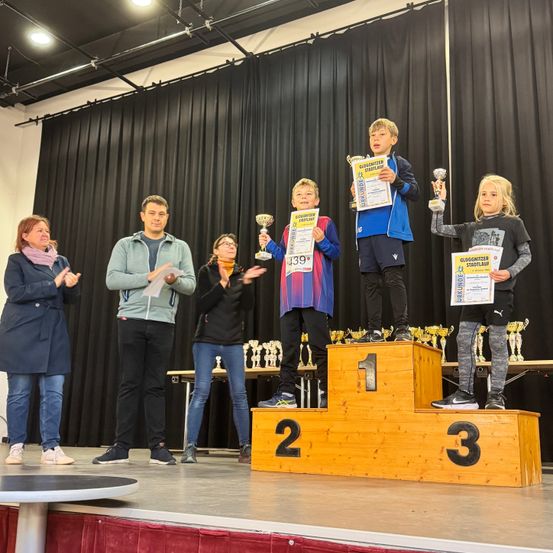 Kinder halten Trophäen und stehen auf einem Podest mit den Nummern 1, 2 und 3, während Erwachsene zusehen. Die Bühne ist mit Trophäen und Vorhängen geschmückt.