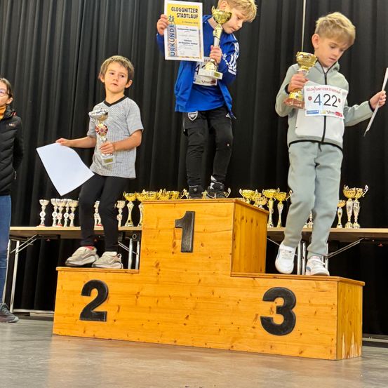 Drei Jungen stehen auf einem Podest und halten Pokale. Ein Junge hält ein Zertifikat, ein anderer ein Papier. Das Podest hat die Zahlen 1, 2 und 3. Dahinter steht ein Tisch mit Pokalen und einem schwarzen Vorhang.