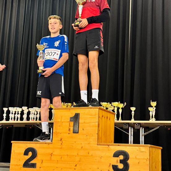 Zwei Jungen stehen auf einem Holzpodium, einer hält einen Pokal. Der Junge auf der rechten Seite trägt ein rotes Shirt und schwarze Shorts. Der Junge auf der linken Seite trägt ein blaues Shirt und schwarze Shorts. Hinter ihnen stehen mehrere Trophäen und ein schwarzer Hintergrund.