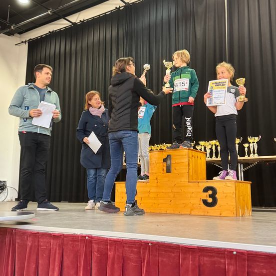 Auf einer Bühne halten drei Kinder Pokale in den Händen, während sie auf einem Podest stehen. Eine Frau reicht ihnen die Pokale. Ein Mann steht neben ihnen mit einem Papier in der Hand. Hinter ihnen steht eine Frau und ein Tisch mit Pokalen im Hintergrund.