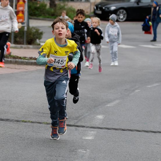 Ein Junge mit der Nummer 145 auf seinem Shirt läuft in einem Rennen mit anderen Kindern. Hinter ihm laufen auch einige Leute. Einige Leute stehen am Straßenrand, und ein Auto ist in der Ferne geparkt.