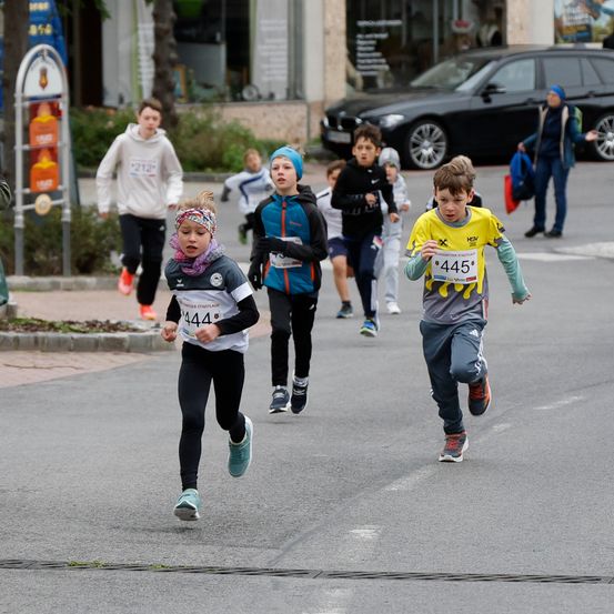 Kinder mit Startnummern laufen in einem Straßenrennen. Sie sind von Gebäuden, einem geparkten Auto und einer Person mit einer Tasche umgeben.