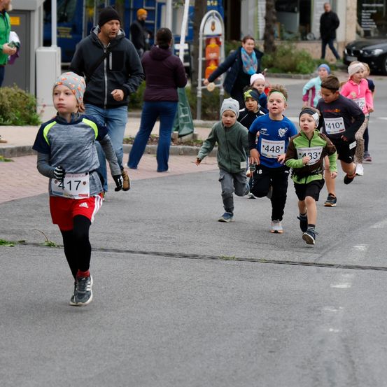 Eine Gruppe von Kindern läuft bei einem Rennen mit Nummern auf ihren Trikots. Zuschauer beobachten von der Seite. Bäume und Gebäude sind im Hintergrund.