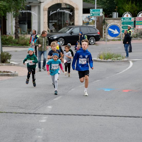 Eine Gruppe von Kindern läuft in einem Rennen, trägt nummerierte Trikots. Sie rennen die Straße hinunter, vorbei an einem Auto und Geschäften. Zuschauer beobachten von der Seite.