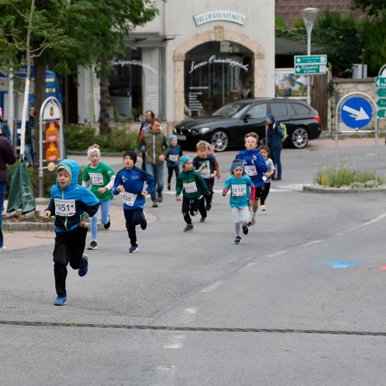 Eine Gruppe von Kindern läuft in einem Rennen auf einer Straße, mit einem Gebäude und einem geparkten Auto im Hintergrund.