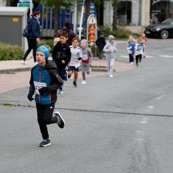 Kinder nehmen an einem Laufwettbewerb auf einer Stadtstraße teil und tragen nummerierte Startnummern. Ein Junge vorne trägt eine blaue Mütze und hat die Nummer 432.