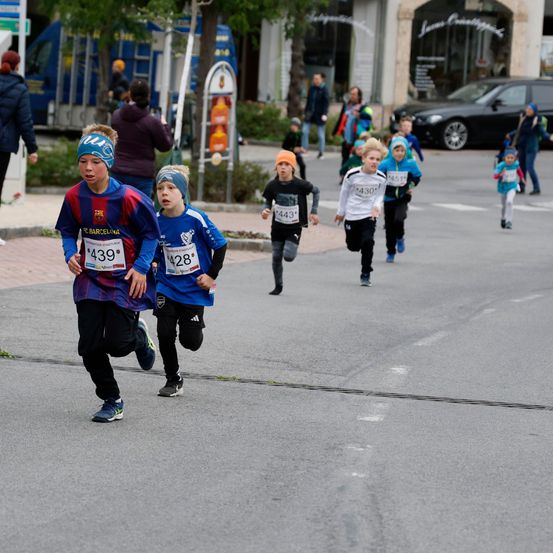 Mehrere Kinder laufen ein Rennen auf der Straße, während einige Leute zusehen. Sie tragen nummerierte Startnummern und Turnschuhe. Dahinter befinden sich Gebäude, ein geparktes Auto und einige weitere Personen.