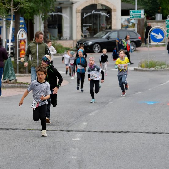 Eine Gruppe von Kindern nimmt an einem Laufevent auf einer Straße teil, an der Geschäfte und geparkte Autos stehen. Ein Mann läuft hinter ihnen.
