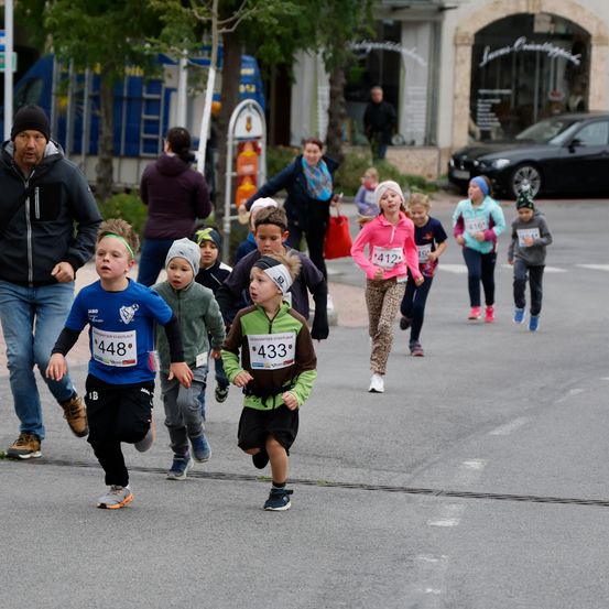 Eine Gruppe von Kindern nimmt an einem Rennen auf der Straße teil. Jedes Kind hat eine Nummer auf seinem Trikot. Zuschauer und Erwachsene gehen in der Nähe. Es gibt Gebäude, ein geparktes Auto und einen Baum im Hintergrund.