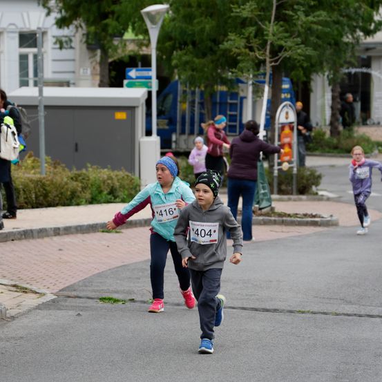 Mehrere Kinder laufen in einem Marathon auf der Straße. Ein Junge mit der Nummer 404 ist in Führung. Hinter ihm läuft ein Mädchen mit der Nummer 416. Andere Leute beobachten.