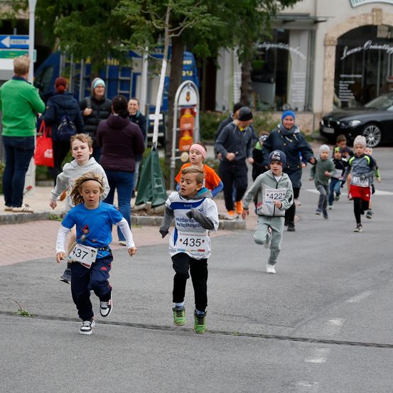 Eine Gruppe von Kindern läuft in einem Rennen, wobei Erwachsene von der Bürgersteigseite zuschauen. Sie tragen nummerierte Startnummern und Turnschuhe. Dahinter stehen und beobachten Menschen, und ein Gebäude ist sichtbar.