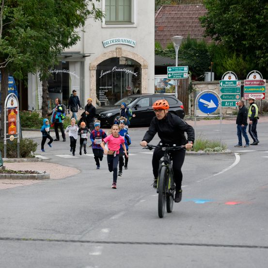 Ein Radfahrer mit orangem Helm fährt durch eine Straße, während Kinder in Laufbekleidung an einem Rennen teilnehmen.