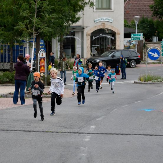 Eine Gruppe von Kindern nimmt an einem Laufevent teil, trägt nummerierte Trikots und Turnschuhe, mit Zuschauern und einem geparkten Auto in der Nähe.