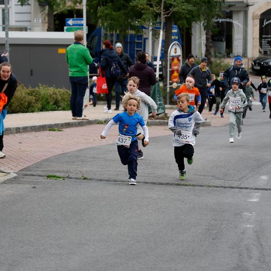 Mehrere Kinder laufen bei einem Rennen auf einer Stadtstraße. Zuschauer beobachten vom Bürgersteig aus.