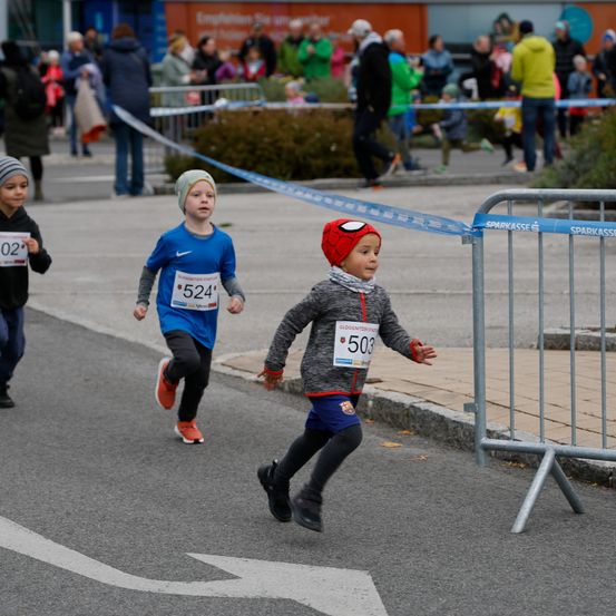 Drei Kinder laufen in einem Rennen. Das vorne hat einen Spider-Man Hut und die Nummer 503 auf seinem Shirt. Dahinter sind zwei andere Kinder, eines trägt ein blaues Shirt mit der Nummer 524. Zuschauer beobachten.