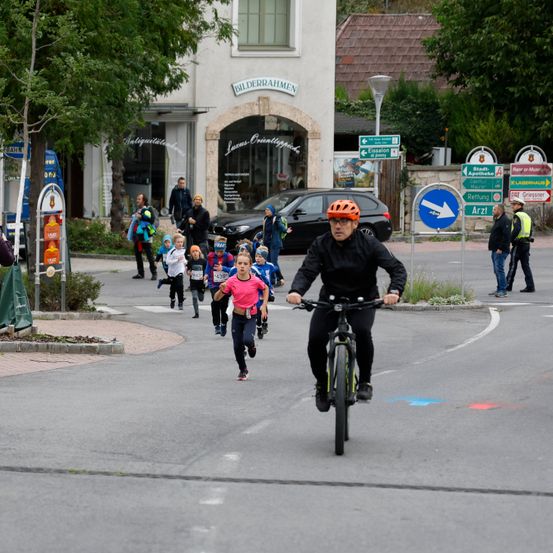 Ein Mann fährt mit einem Fahrrad auf einer Straße, während Kinder ein Rennen laufen. Ein Kind trägt ein pinkes Shirt. Schilder und Straßenlaternen sind im Hintergrund.