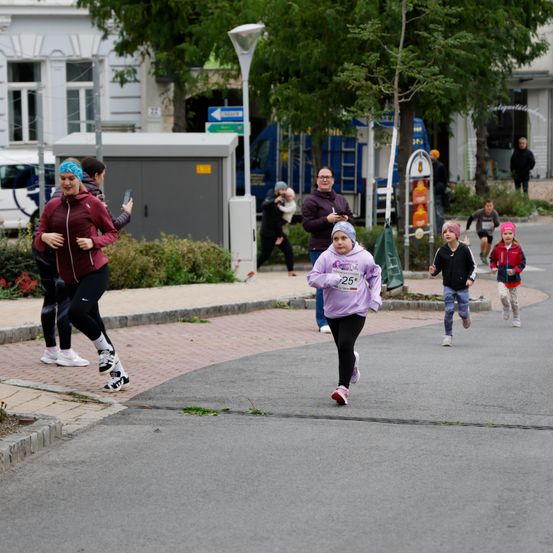 Eine Gruppe von Menschen nimmt an einem Rennen auf einer Straße teil. Ein Mädchen in einem lila Shirt führt, während andere hinterherlaufen. Bäume und ein Gebäude sind im Hintergrund zu sehen.
