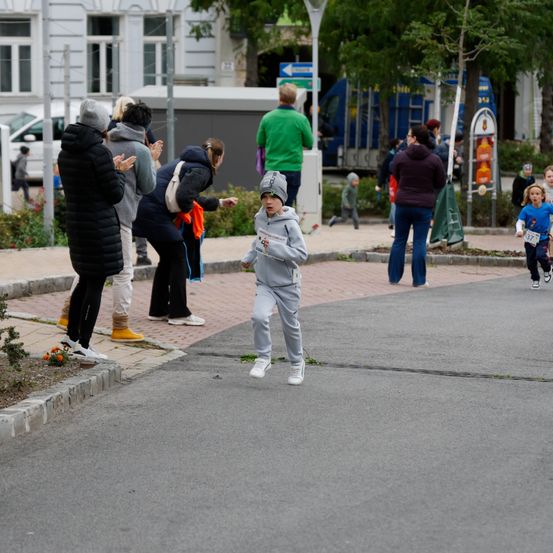 Ein Junge in einer grauen Kapuzenjacke und Turnschuhen läuft auf einer Straße, während einige Leute auf dem Bürgersteig stehen und zusehen.