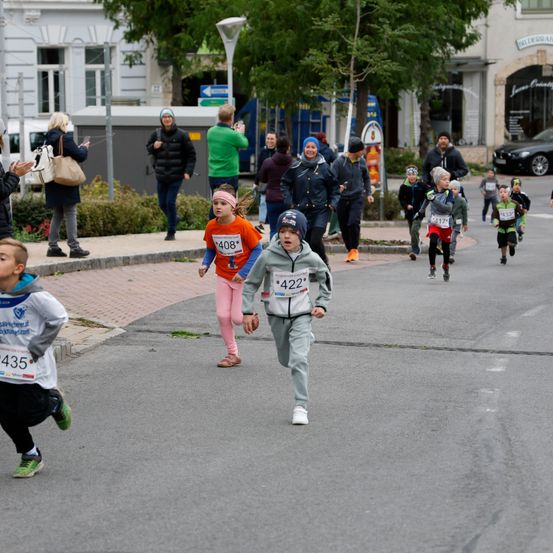 Ein Kinderlaufrennen auf einer Stadtstraße mit Läufern, Zuschauern auf dem Bürgersteig und Gebäuden im Hintergrund.