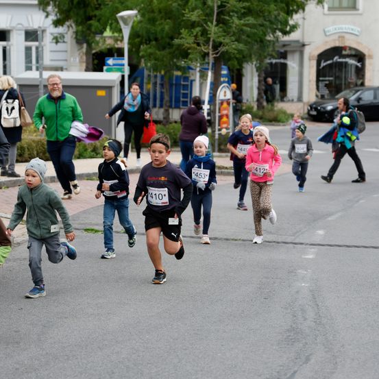 Eine Gruppe von Kindern läuft auf einer Stadtstraße an einem Rennen teil. Im Hintergrund befinden sich Gebäude und Bäume. Zuschauer beobachten sie.