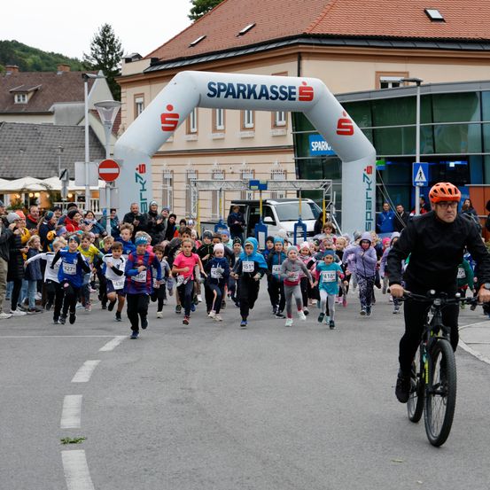 Eine Gruppe von Kindern nimmt an einem Rennen auf einer Straße teil, mit einem Bogen, der das Wort 'Sparkasse' über ihnen anzeigt. Ein Mann fährt in der Nähe ein Fahrrad.