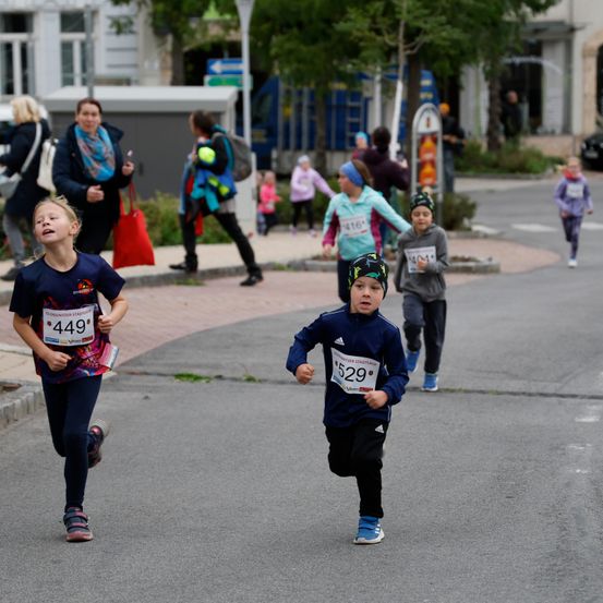 Mehrere Kinder laufen ein Rennen auf einer Straße mit Bäumen und Gebäuden an den Seiten. Ein Junge mit der Nummer 529 führt, während ein Mädchen mit der Nummer 449 hinterherläuft. Zuschauer beobachten vom Bürgersteig.