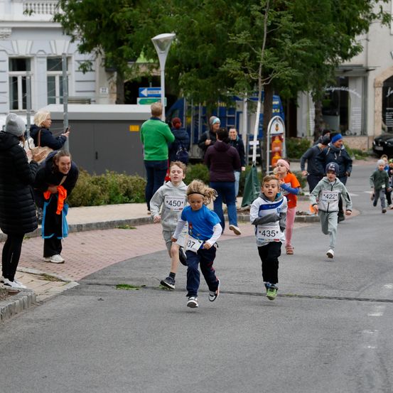 Eine Gruppe von Kindern nimmt an einem Rennen auf einer Stadtstraße teil. Zuschauer beobachten vom Bürgersteig aus. Bäume und Gebäude säumen die Straße.