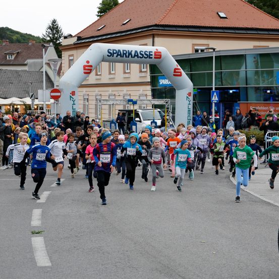 Eine große Gruppe von Kindern läuft auf einer Straße mit Zuschauern und einem Gebäude mit dem Wort 'Sparkasse'.