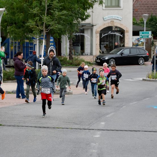 Eine Gruppe von Kindern nimmt an einem Rennen in einer Stadt teil. Sie laufen auf einer Straße, und einige Leute stehen auf dem Bürgersteig. Ein schwarzer Wagen ist auf der Straße geparkt. Bäume und Straßenlaternen befinden sich an der Seite. Ein Gebäude mit einem Schild ist rechts.