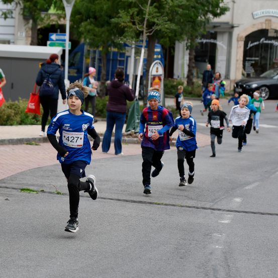Mehrere Kinder laufen bei einem Rennen auf der Straße. Eines von ihnen trägt ein blaues Shirt mit der Nummer 427. Menschen stehen auf dem Bürgersteig.