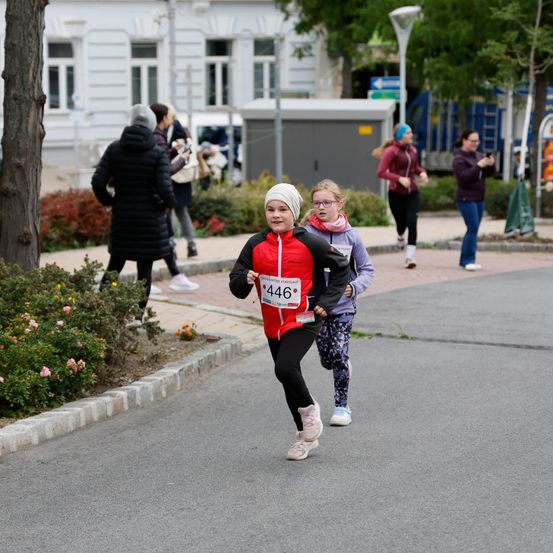 Eine Gruppe von Läufern nimmt an einem Rennen auf einer Stadtstraße teil. Ein Mädchen in einer rot-schwarzen Jacke mit der Nummer 446 führt die Gruppe an.