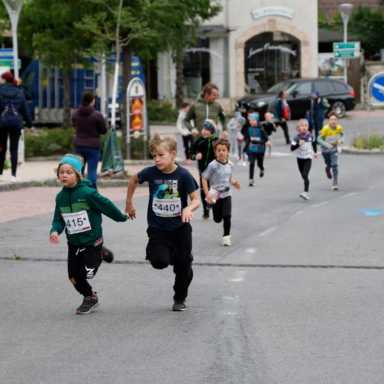 Mehrere Kinder nehmen an einem Rennen auf einer Straße teil, mit Nummern auf ihren Trikots. Ein Mann läuft hinter ihnen. Im Hintergrund befinden sich Gebäude, Bäume und geparkte Autos.