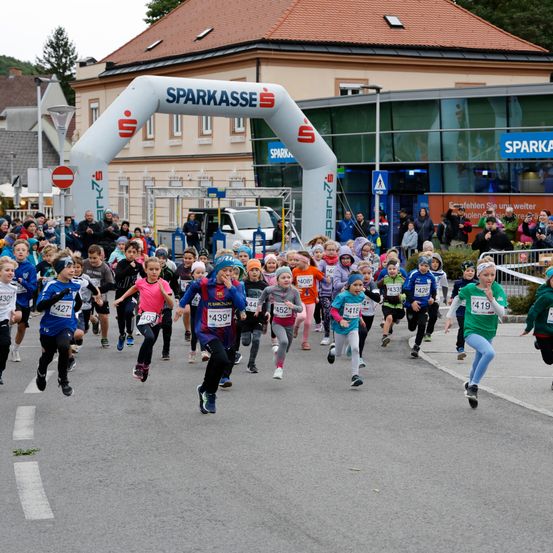 Eine Gruppe von Kindern mit nummerierten Startnummern läuft einen Wettkampf auf einem offenen Gelände. Ein großer Bogen mit der Aufschrift 'Sparkasse' steht im Hintergrund. Zuschauer beobachten von der Straßenseite.