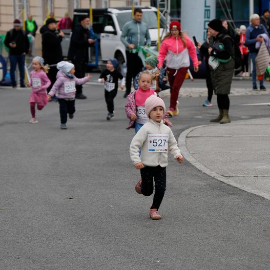 Eine Gruppe von Kindern bei einem Rennen, mit der Nummer 527 vorne, in einer weißen Jacke und einem rosa Hut. Zuschauer stehen am Straßenrand.
